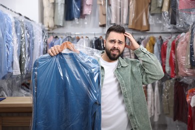 Photo of Dry-cleaning service. Confused man holding hanger with sweatshirt in plastic bag indoors