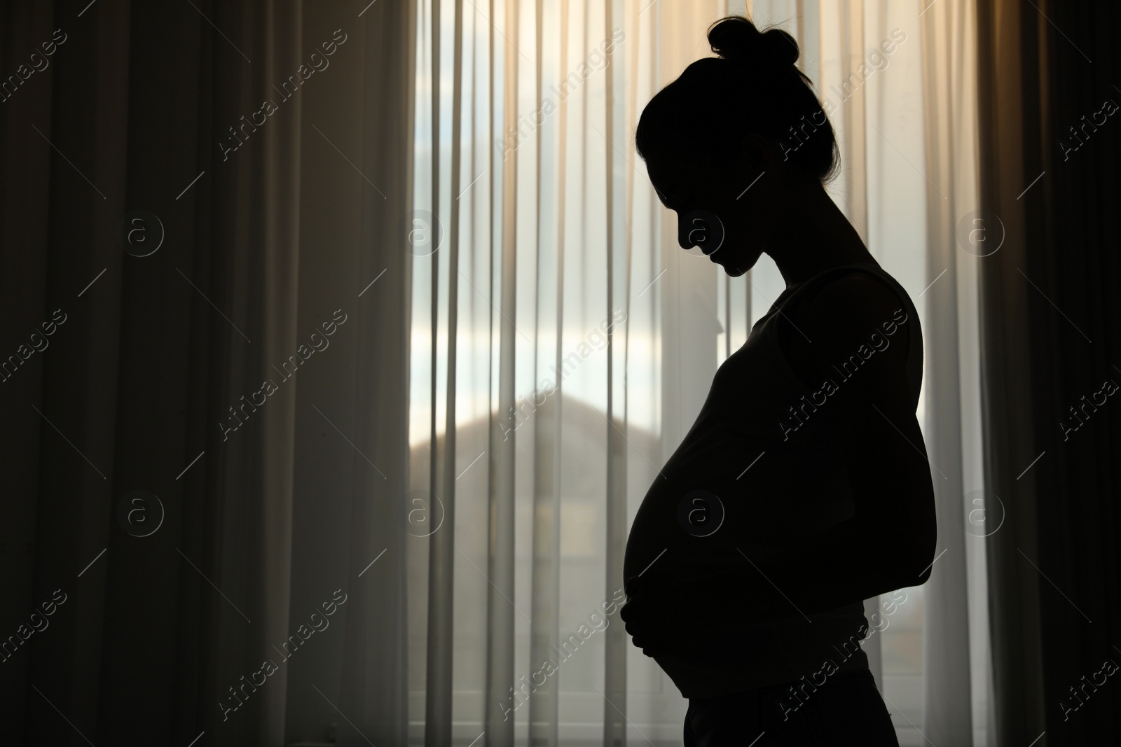 Photo of Silhouette of young pregnant woman near window at home. Space for text