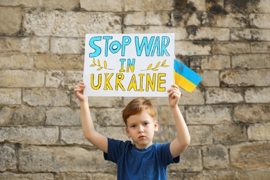 Photo of Sad boy holding poster in colors of national flag with words Stop War In Ukraine against brick wall outdoors