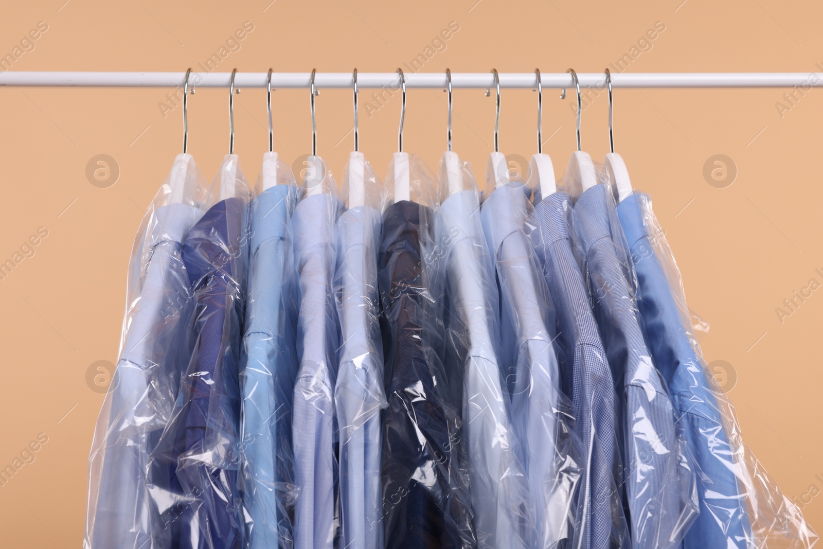 Photo of Dry-cleaning service. Many different clothes in plastic bags hanging on rack against beige background