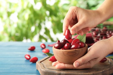 Photo of Woman taking fresh ripe dogwood berry from bowl at light blue wooden table, closeup. Space for text