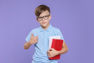 Photo of Cute schoolboy in glasses holding books and showing thumbs up on violet background