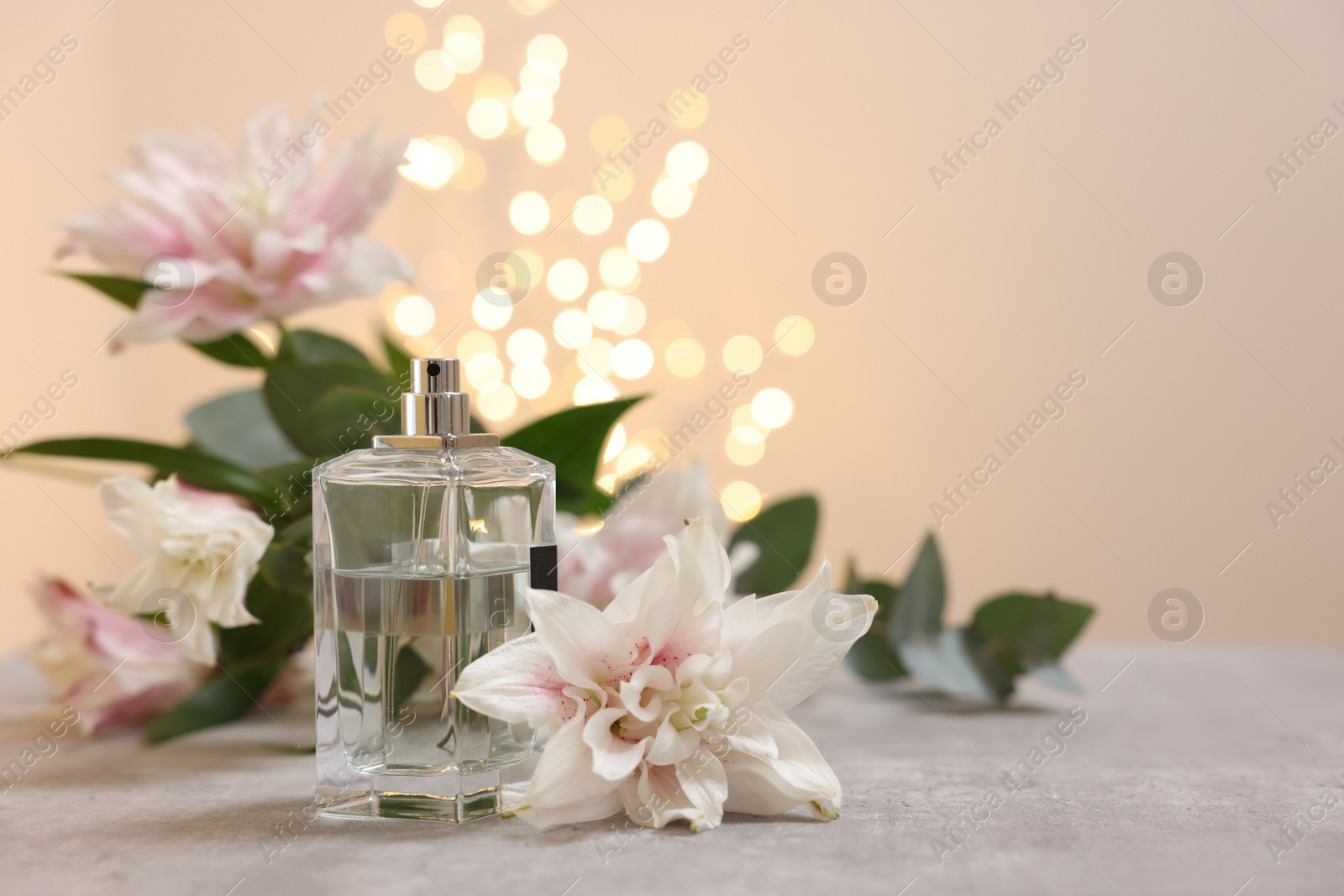 Photo of Bottle of perfume and beautiful lily flowers on table against beige background with blurred lights, space for text