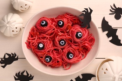 Photo of Red pasta with decorative eyes and olives in bowl served on white wooden table, flat lay. Halloween food