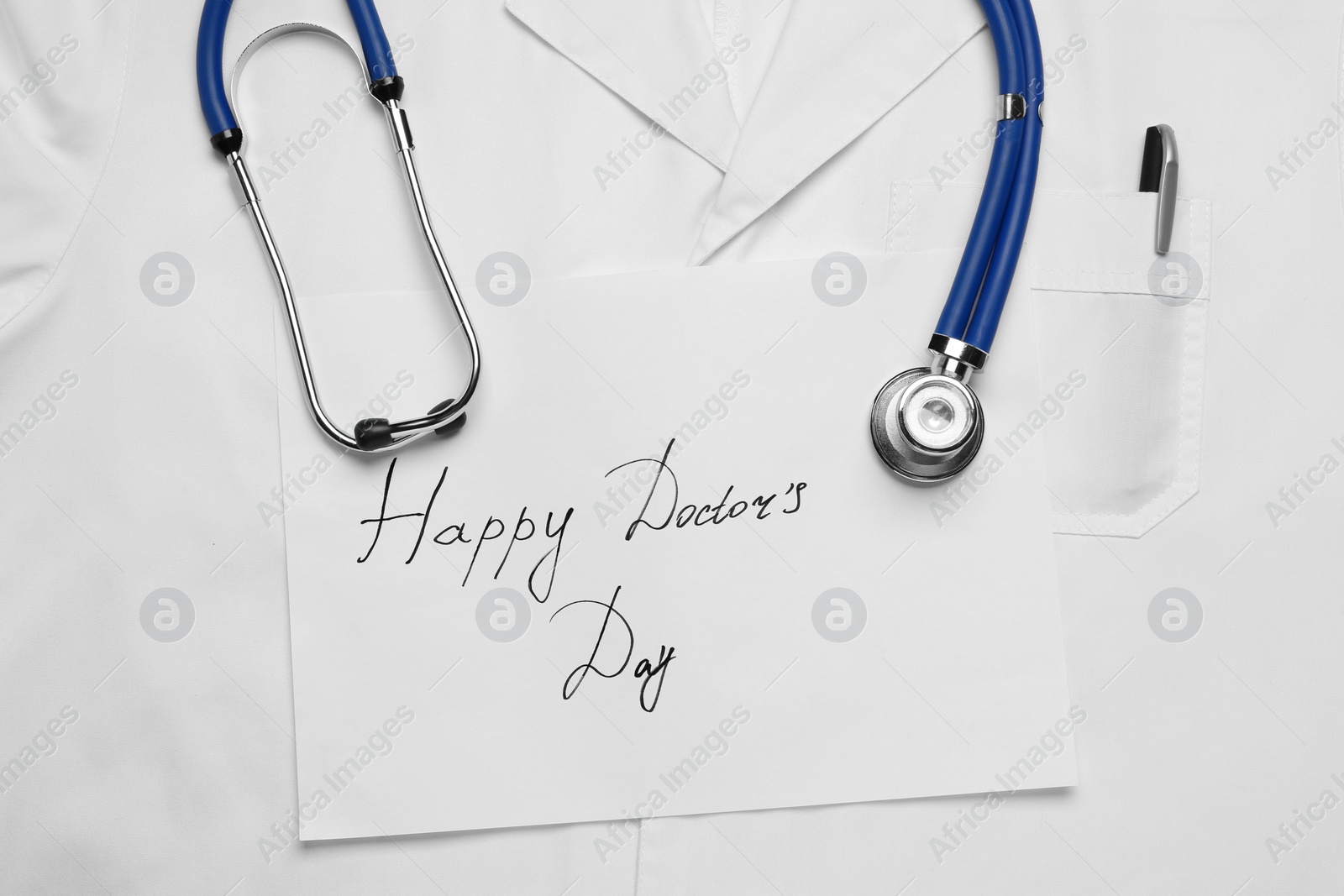 Photo of Stethoscope and paper sheet with phrase Happy Doctor's Day on white medical uniform, top view