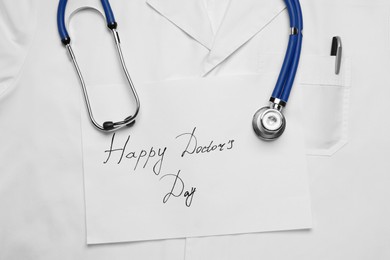 Stethoscope and paper sheet with phrase Happy Doctor's Day on white medical uniform, top view