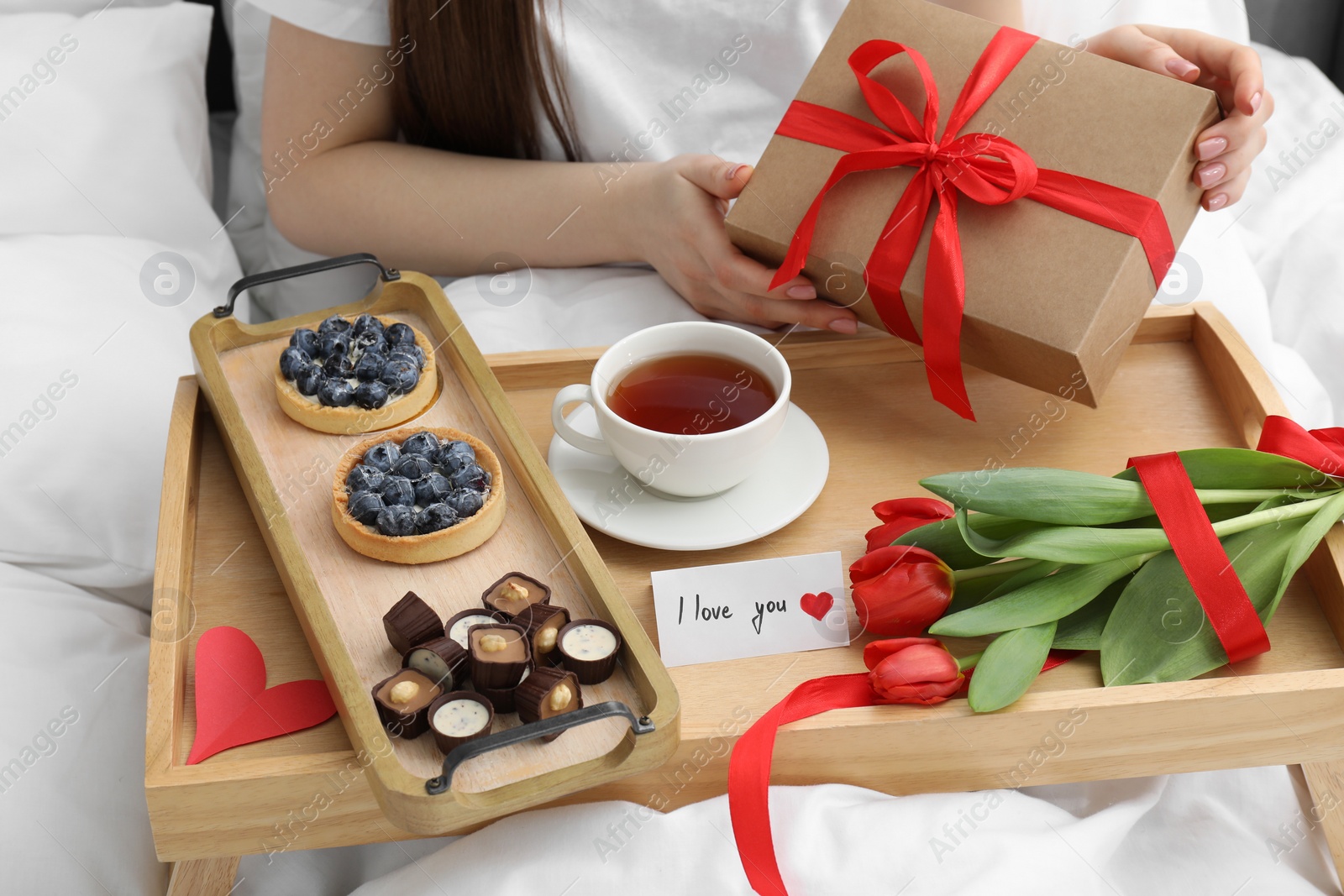 Photo of Tasty breakfast served in bed. Woman with gift box, desserts, tea, flowers and I Love You card at home, closeup