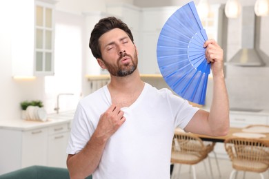 Photo of Bearded man waving blue hand fan to cool himself in kitchen