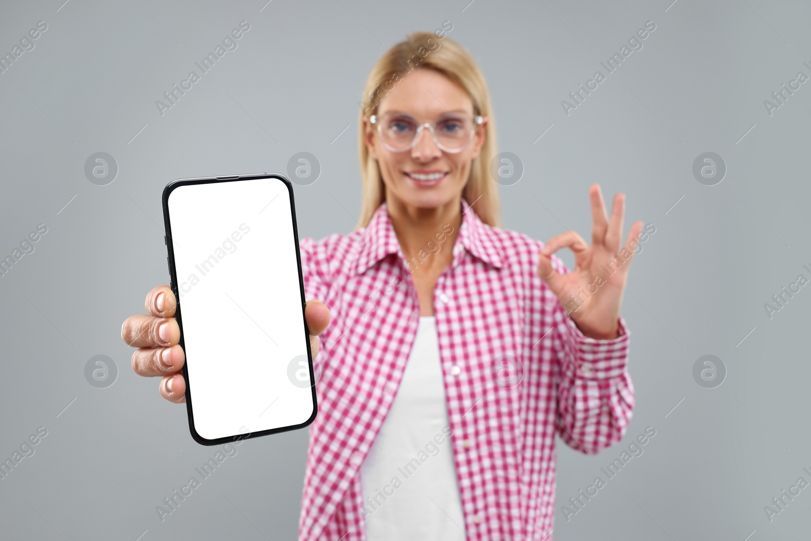 Photo of Happy woman holding smartphone with blank screen and showing OK gesture on grey background, selective focus