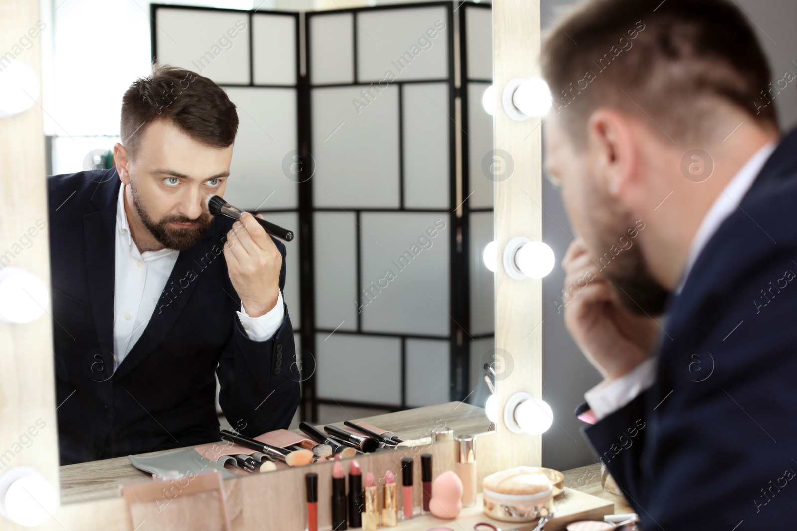 Photo of Young man using makeup brush near mirror in dressing room