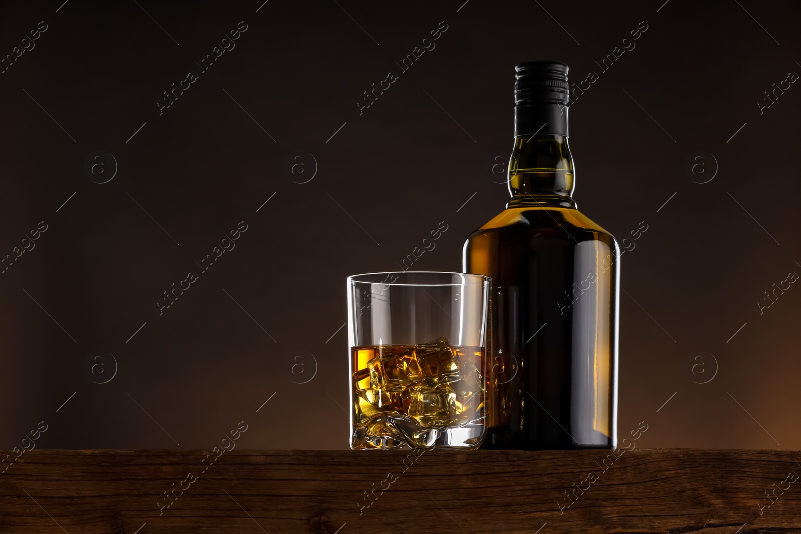 Photo of Whiskey with ice cubes in glass and bottle on wooden table, space for text