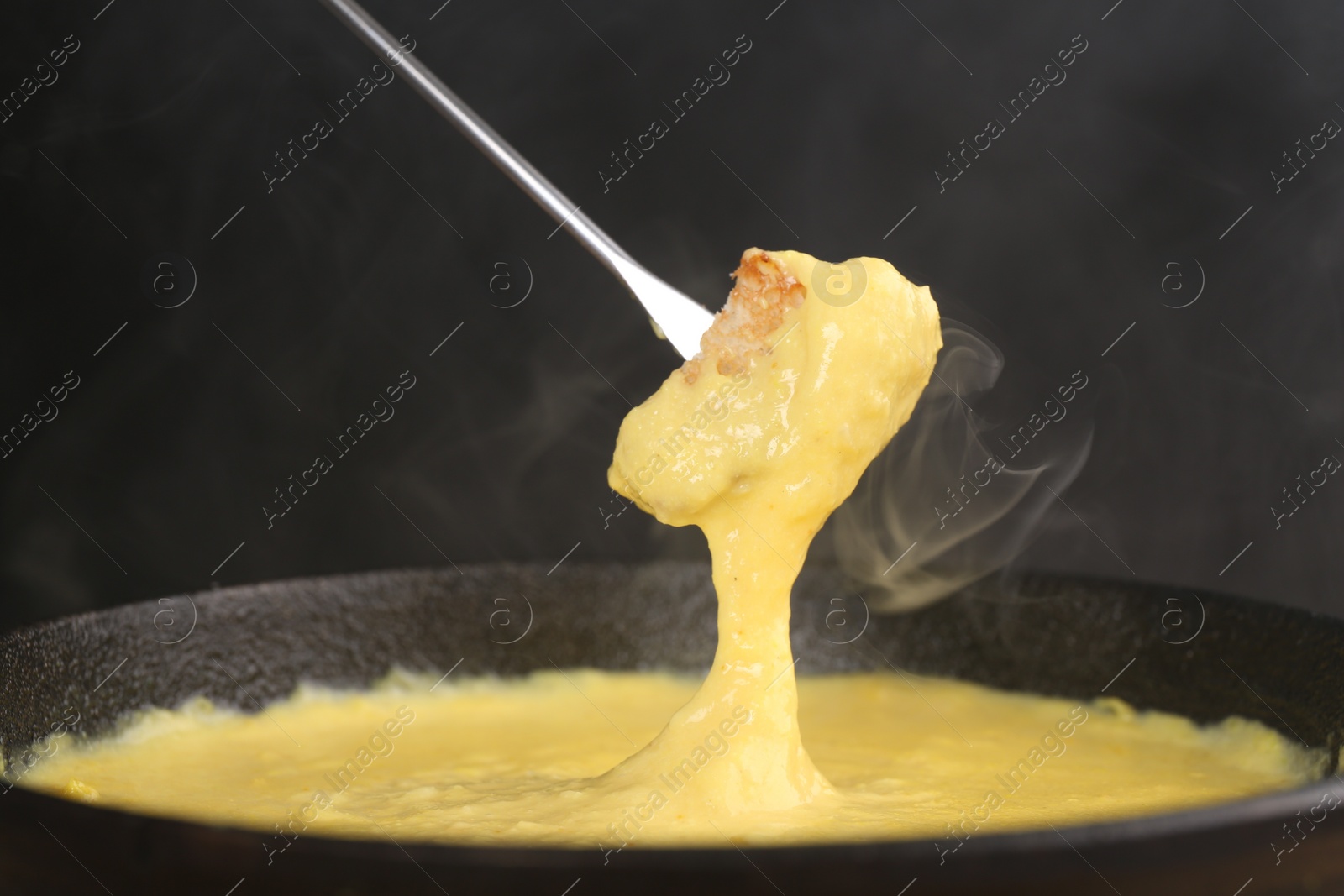 Photo of Dipping piece of bread into fondue pot with tasty melted cheese against dark gray background, closeup