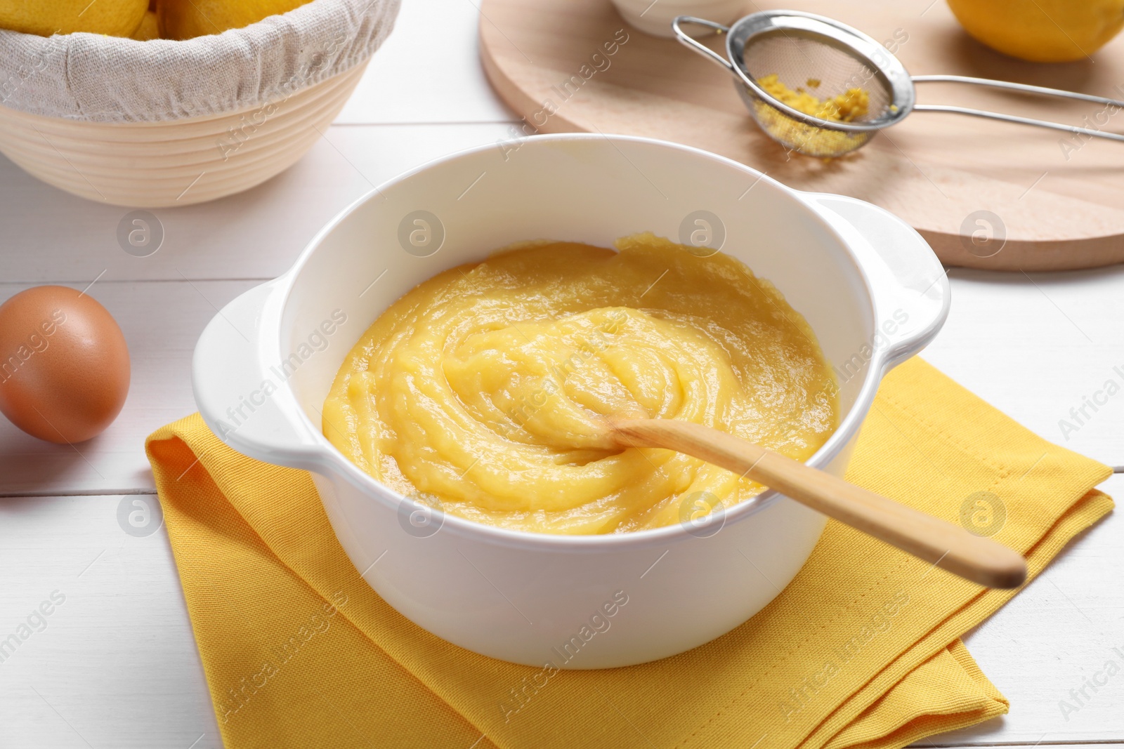 Photo of Delicious lemon curd in bowl, ingredients, spoon and sieve on white wooden table, closeup