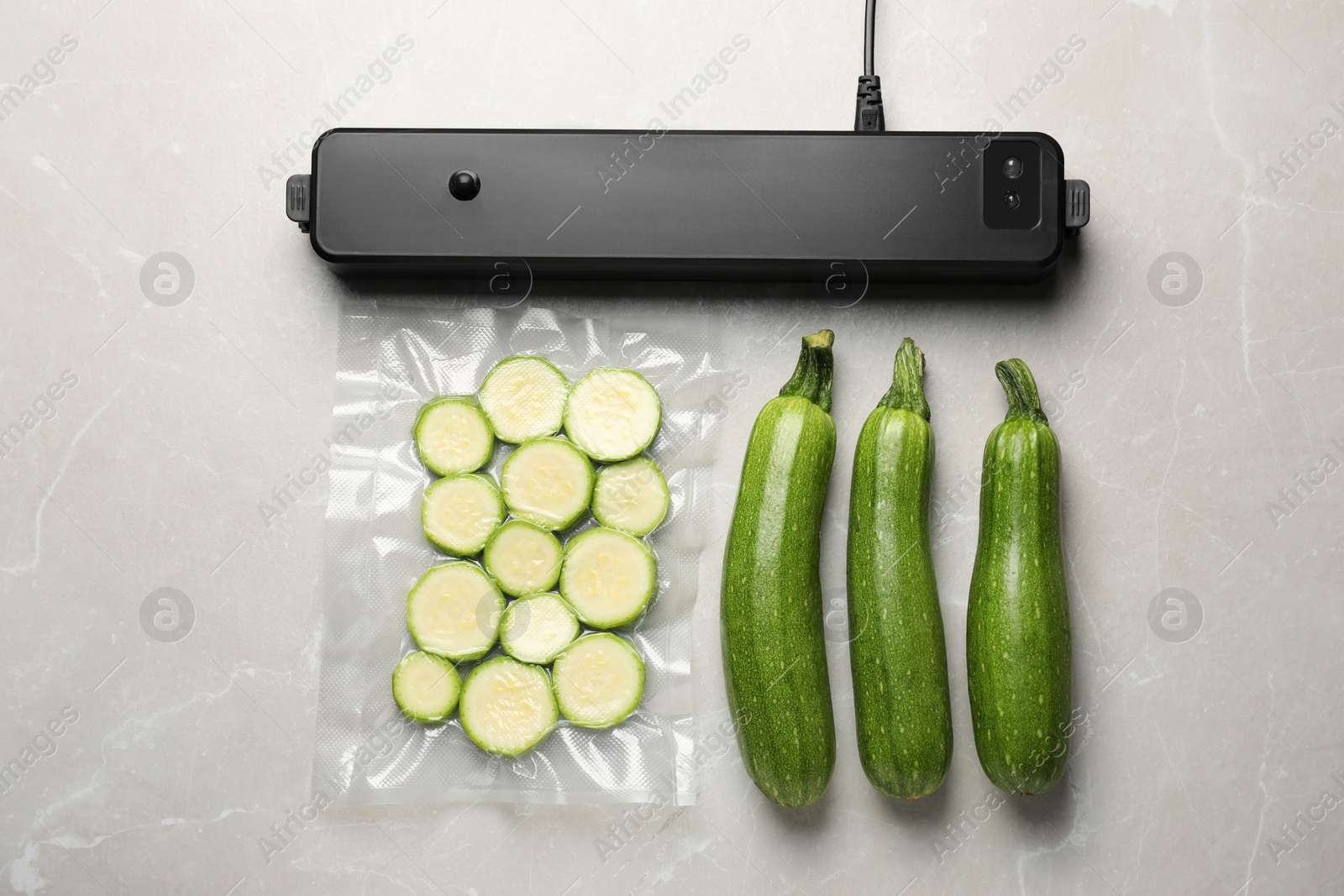 Photo of Sealer for vacuum packing, plastic bag with cut zucchinis and whole vegetables on light grey marble table, flat lay