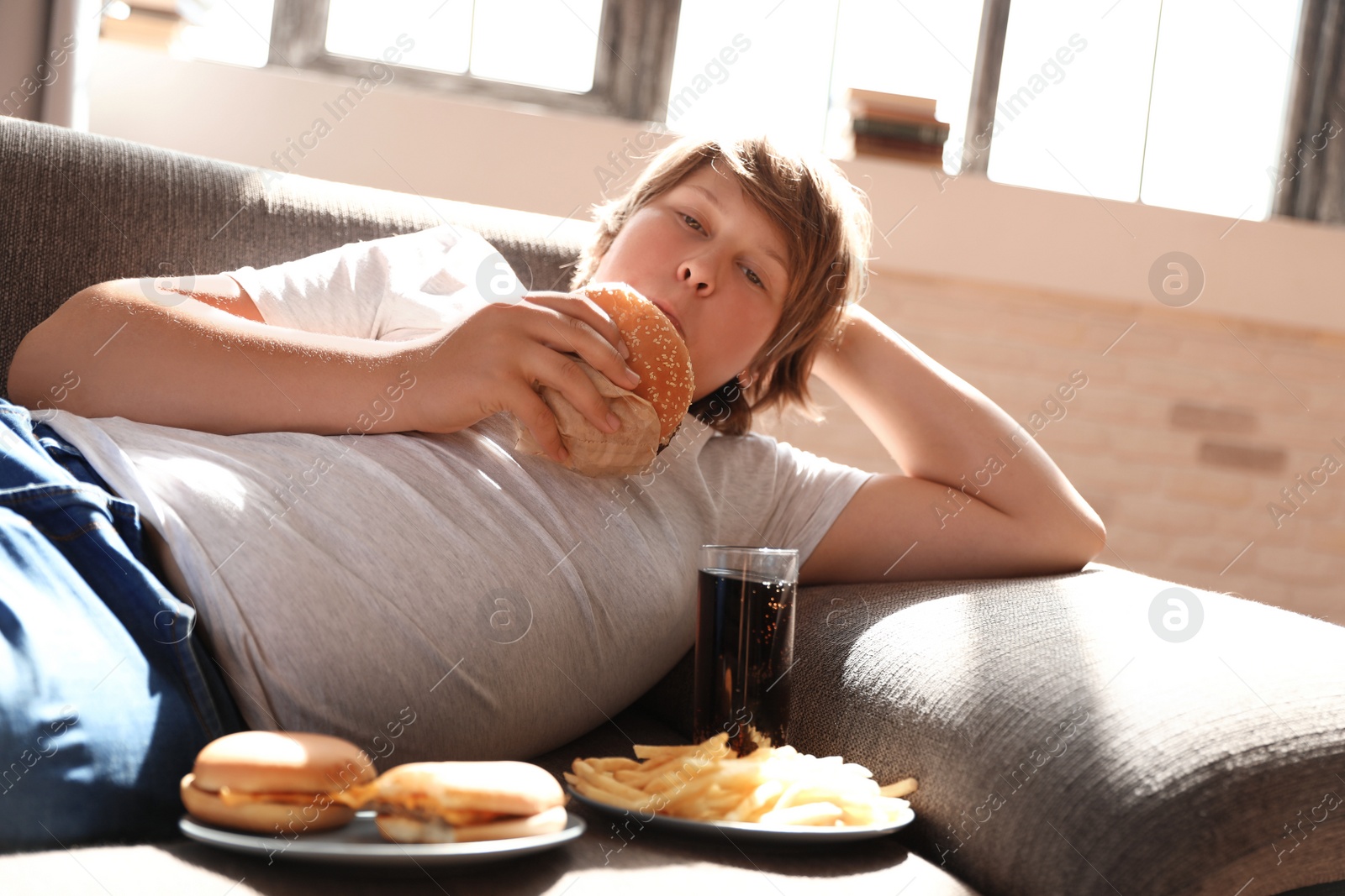 Photo of Overweight boy with fast food on sofa at home