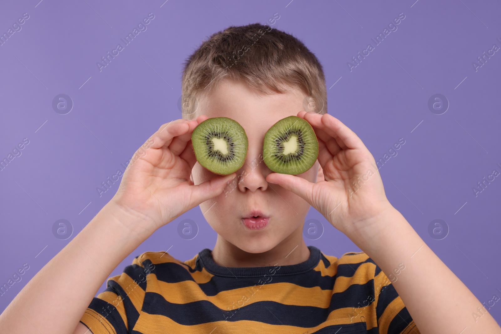 Photo of Emotional boy covering eyes with halves of fresh kiwi on violet background