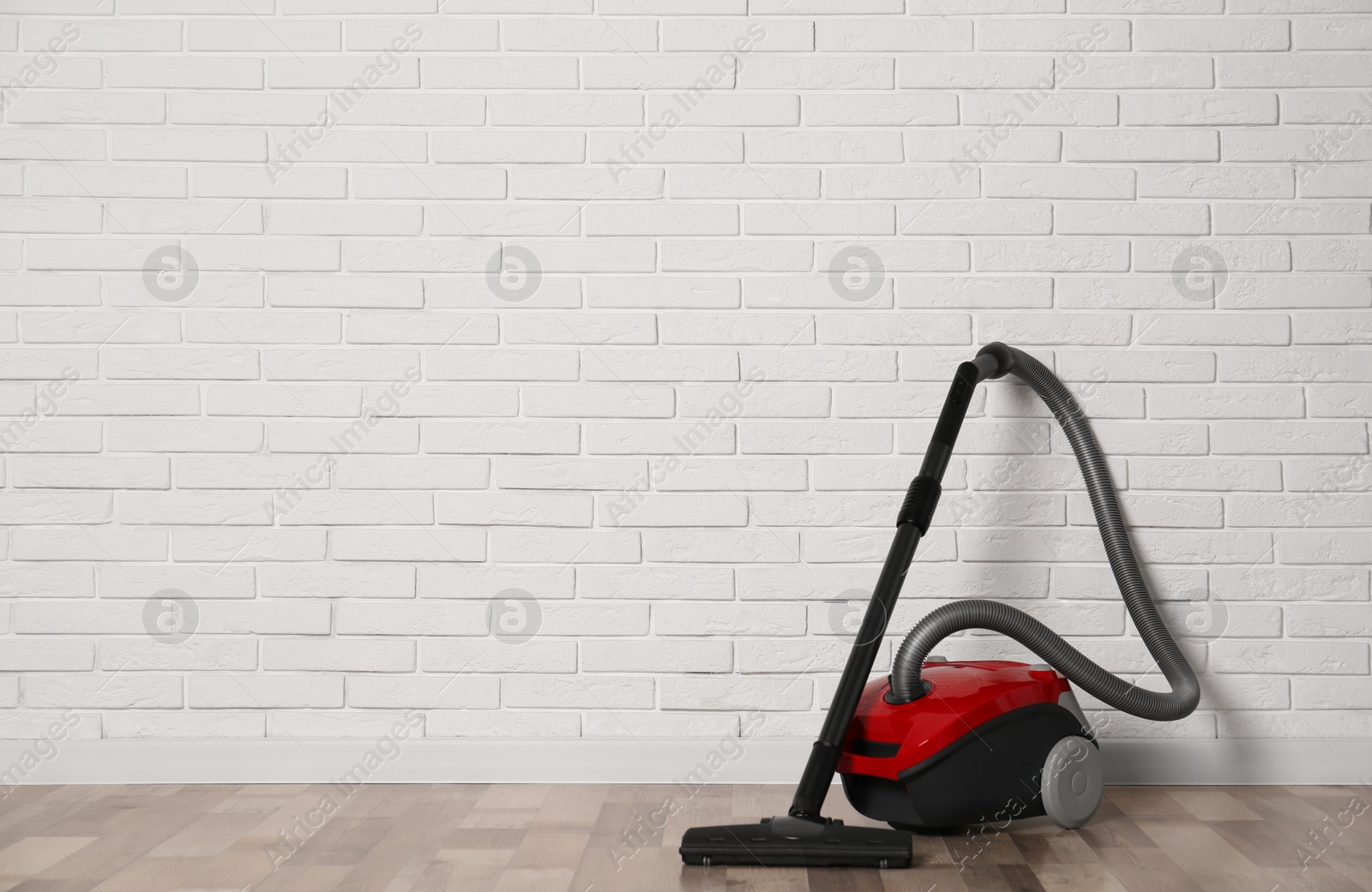 Photo of Modern red vacuum cleaner on wooden floor near white brick wall, space for text