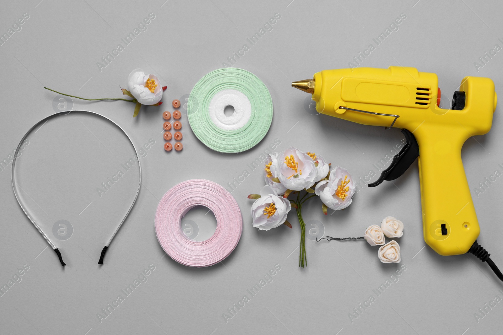 Photo of Hot glue gun, plastic headband and handicraft materials on grey background, flat lay