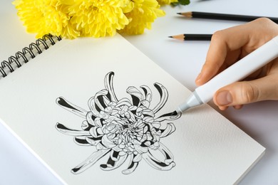 Photo of Woman drawing chrysanthemum in sketchbook at white table, closeup