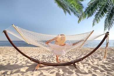 Image of Woman relaxing in hammock under green palm leaves on sunlit beach