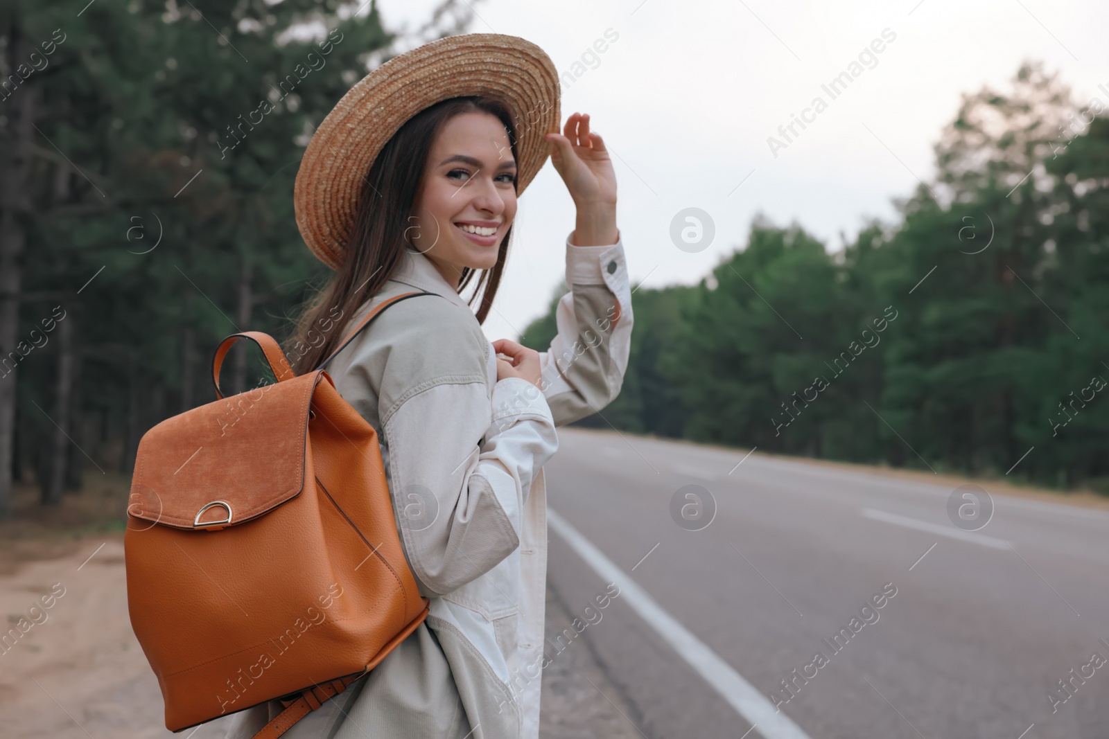 Photo of Young woman with stylish backpack near empty road in forest. Space for text