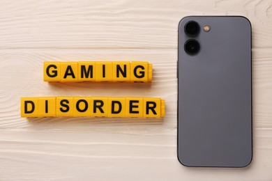 Photo of Phrase Gaming Disorder made of yellow cubes and smartphone on white wooden table, flat lay. Addictive behavior