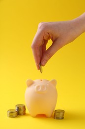 Photo of Financial savings. Woman putting coin into piggy bank on yellow background, closeup