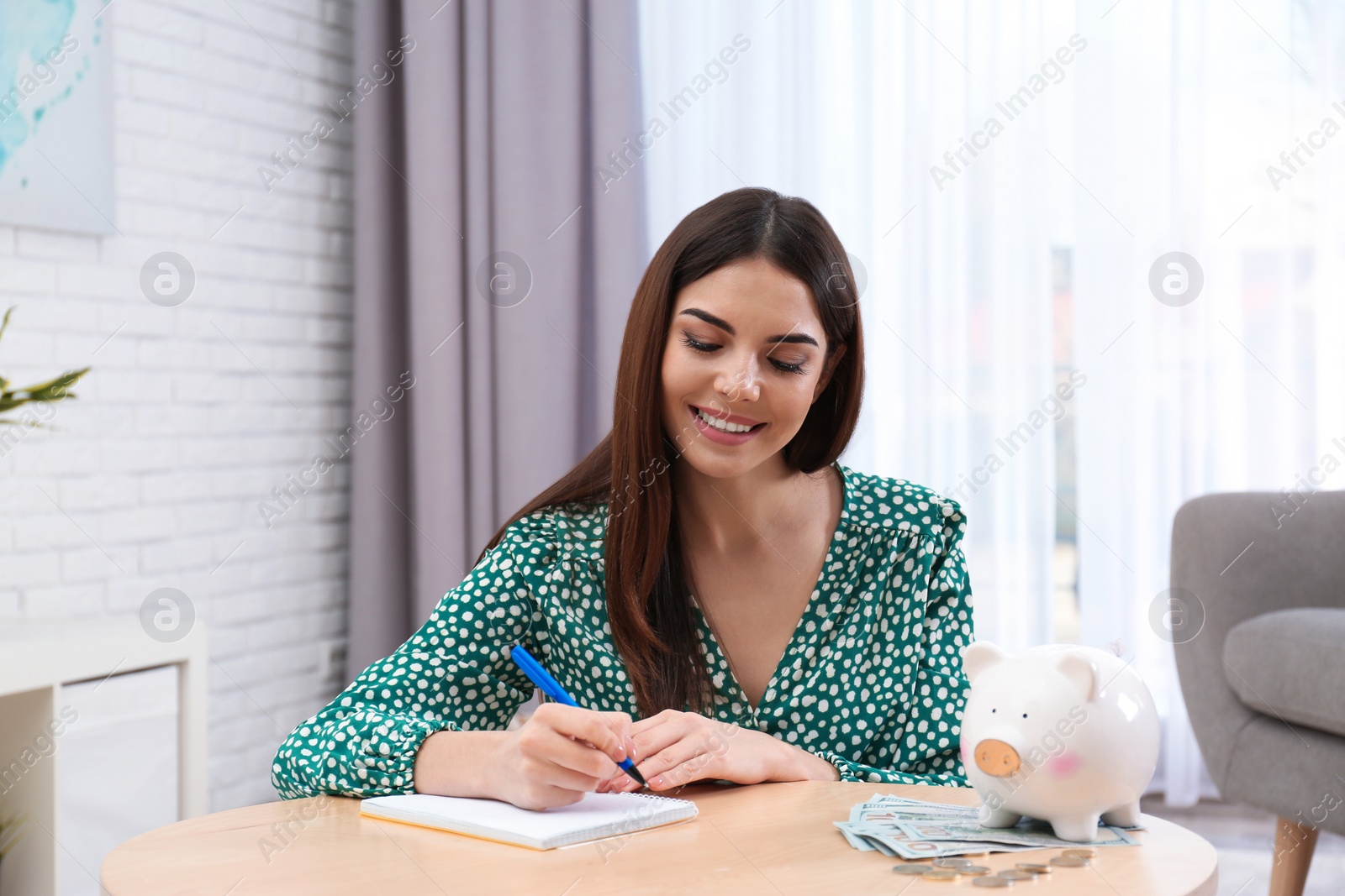 Photo of Happy young woman with piggy bank and money at home