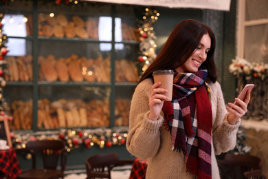 Photo of Beautiful woman with cup of coffee using mobile phone in decorated cafe. Christmas celebration