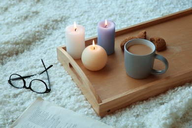 Eyeglasses near wooden tray with burning candles, coffee and cookies on soft fabric