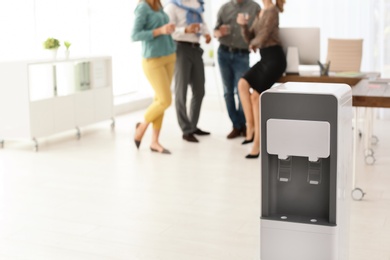 Photo of Modern water cooler and blurred office employees on background. Space for text
