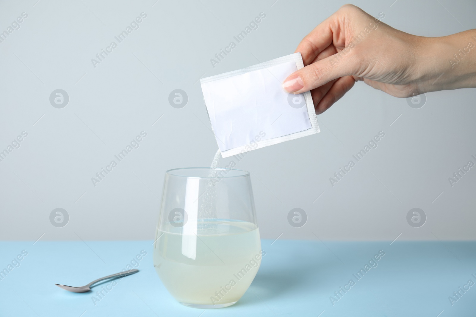Photo of Woman pouring powder from medicine sachet into glass of water on turquoise table, closeup