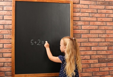 Photo of Cute little left-handed girl doing sums on chalkboard near brick wall