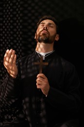 Photo of Catholic priest in cassock holding cross and praying to God in confessional