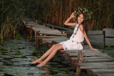 Photo of Cute little girl wearing wreath made of beautiful flowers on pier near pond at sunset