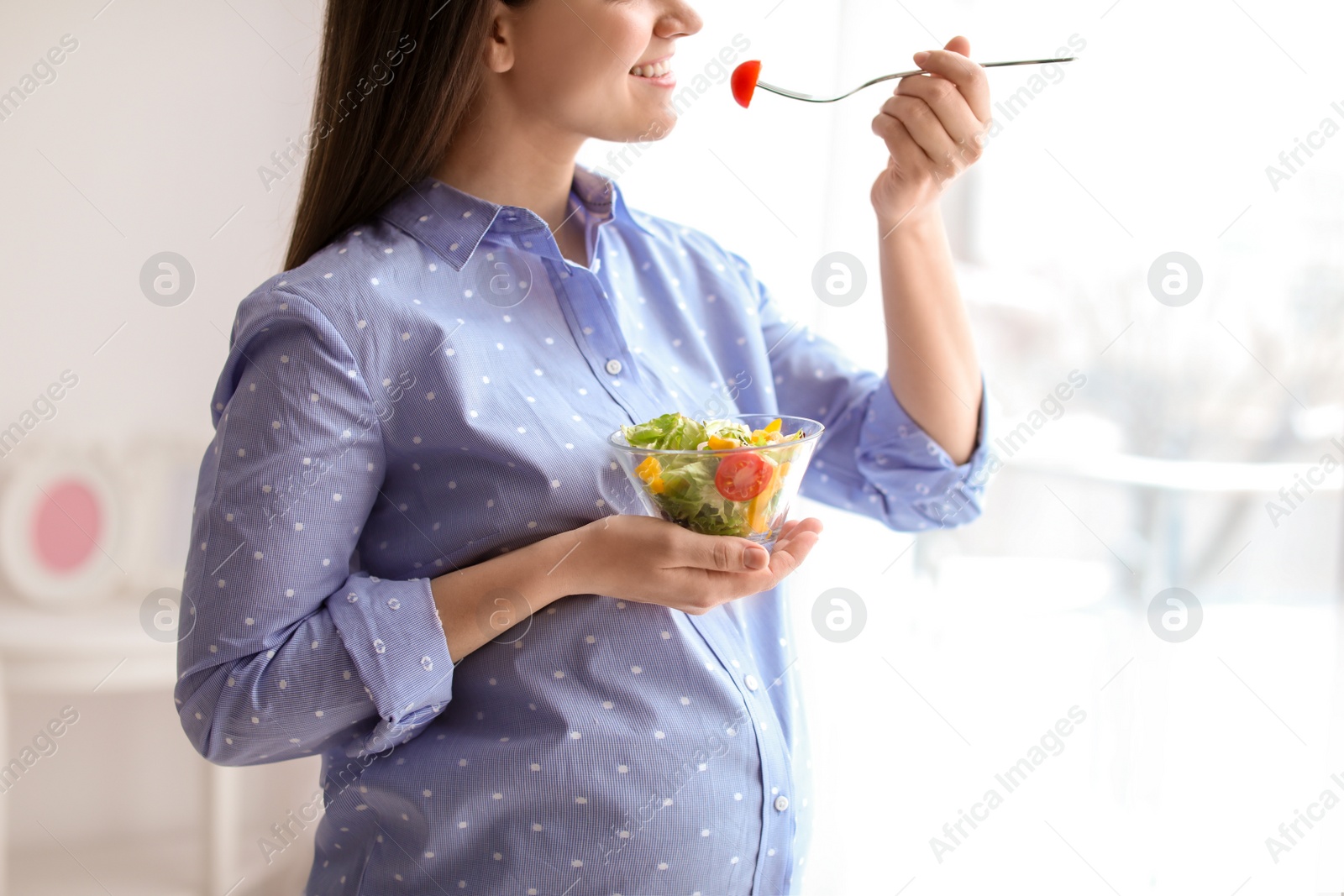 Photo of Young pregnant woman eating vegetable salad near window at home