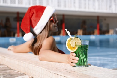 Photo of Young woman wearing Santa Claus hat with refreshing drink in swimming pool. Christmas vacation