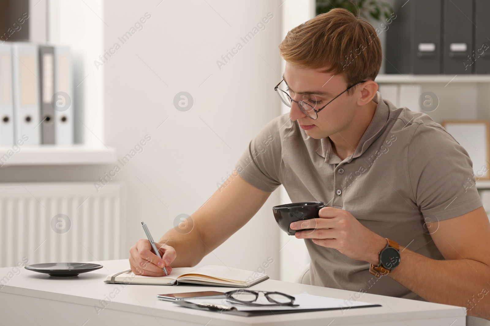 Photo of Young man with cup of coffee writing in notebook at table indoors