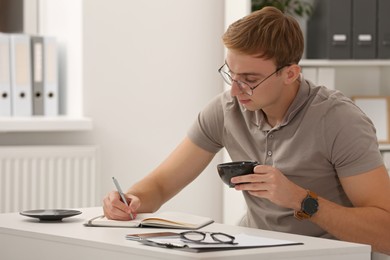 Young man with cup of coffee writing in notebook at table indoors