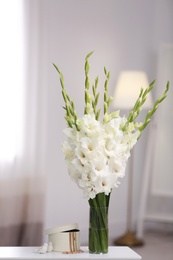 Vase with beautiful white gladiolus flowers and jewellery on wooden table in room, space for text