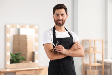 Photo of Smiling hairdresser in apron holding scissors near vanity mirror in salon