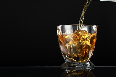 Pouring whiskey into glass with ice cubes at table against black background, space for text