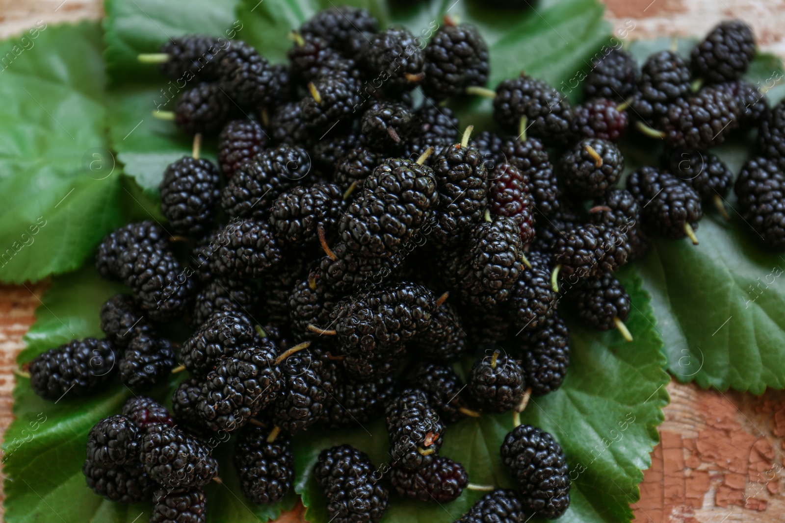 Photo of Heap of delicious ripe black mulberries and green leaves on wooden table, closeup