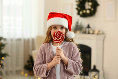 Photo of Cute little girl in Santa hat with lollipop near fireplace at home. Christmas atmosphere
