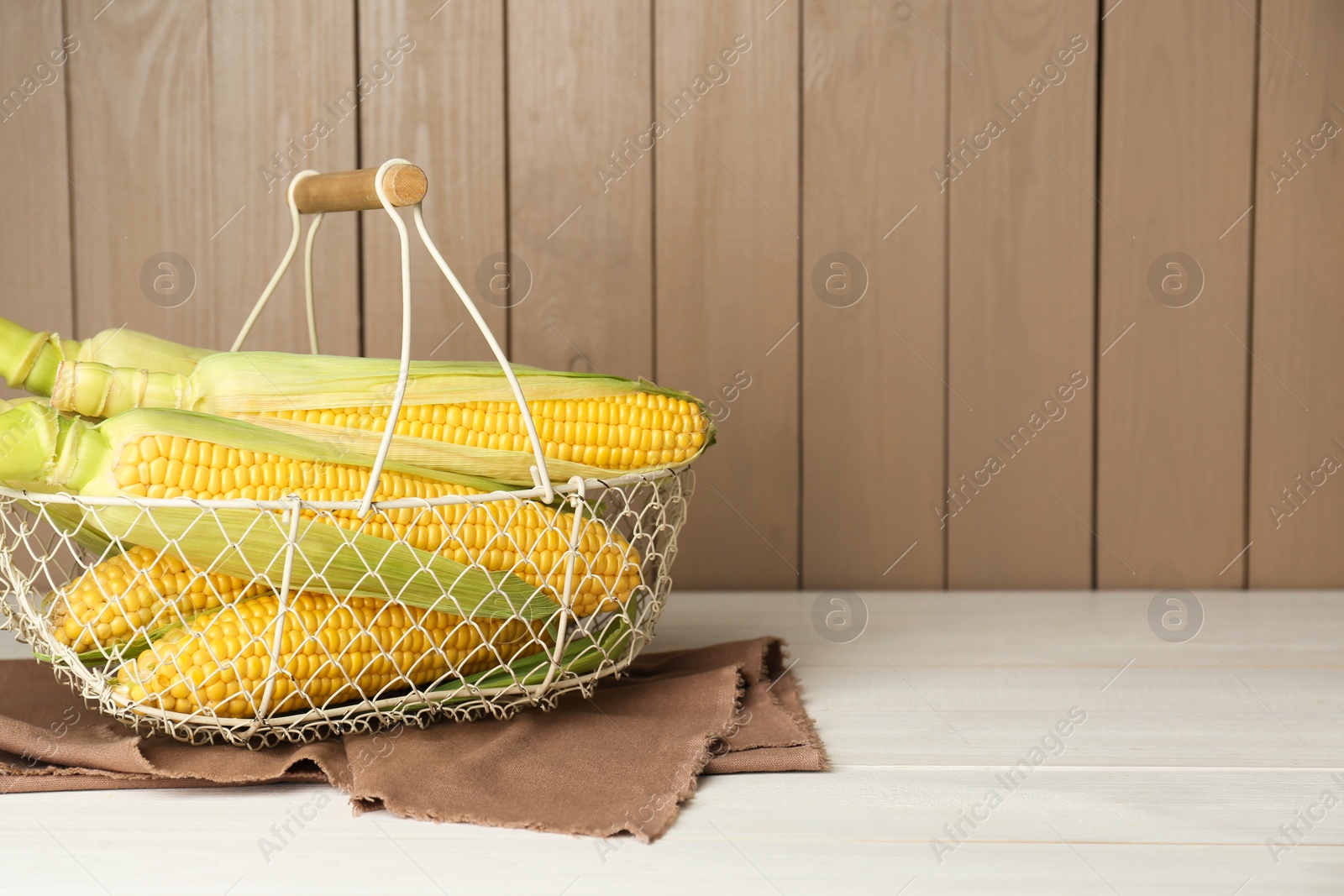 Photo of Tasty fresh corn cobs in metal basket on white wooden table, space for text