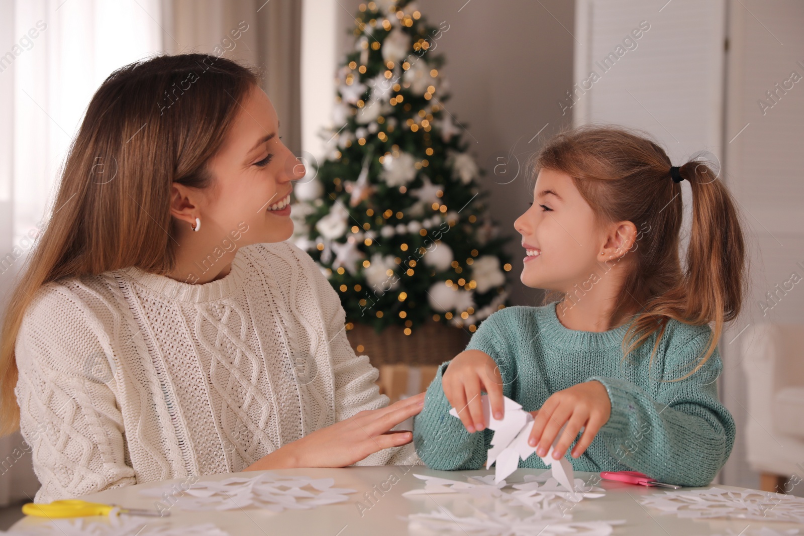 Photo of Happy mother and daughter making paper snowflake at table near Christmas tree indoors