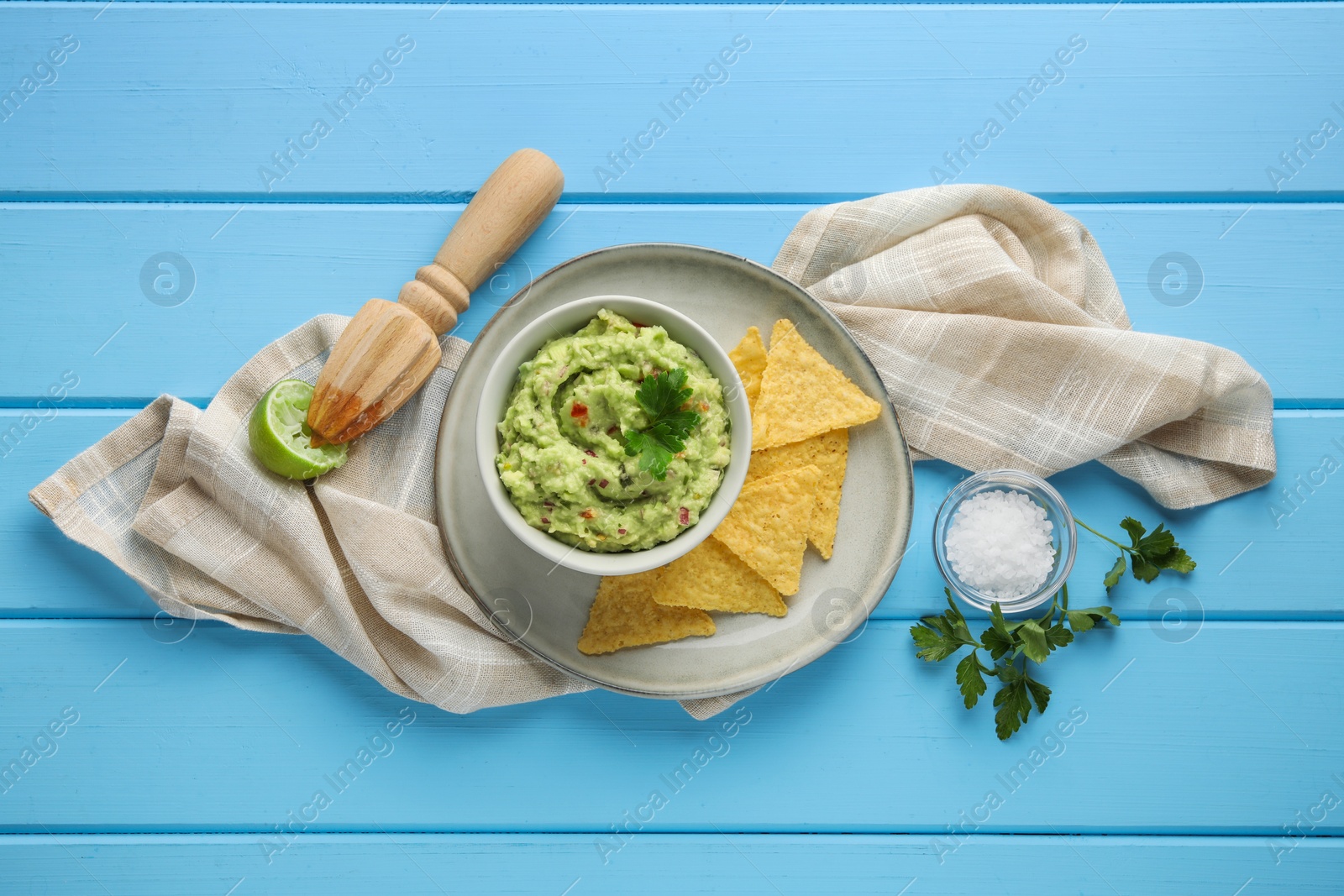 Photo of Delicious guacamole with nachos chips and ingredients on light blue wooden table, flat lay