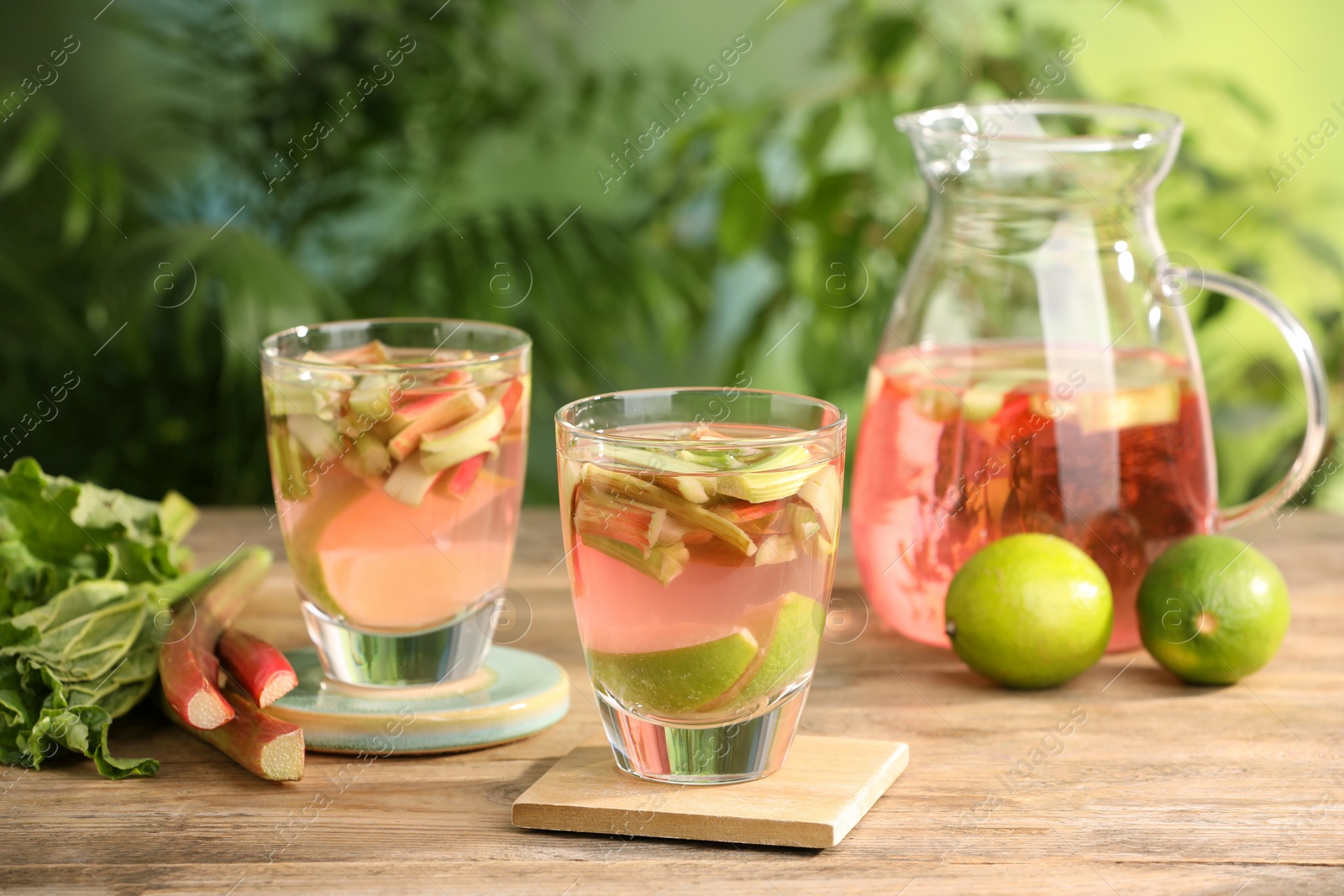 Photo of Glasses and jug of tasty rhubarb cocktail with lime fruits on wooden table outdoors