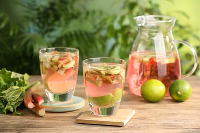 Photo of Glasses and jug of tasty rhubarb cocktail with lime fruits on wooden table outdoors