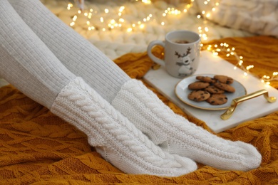 Woman in warm knitted socks sitting near tray with cup of hot drink and cookies on floor at home, closeup